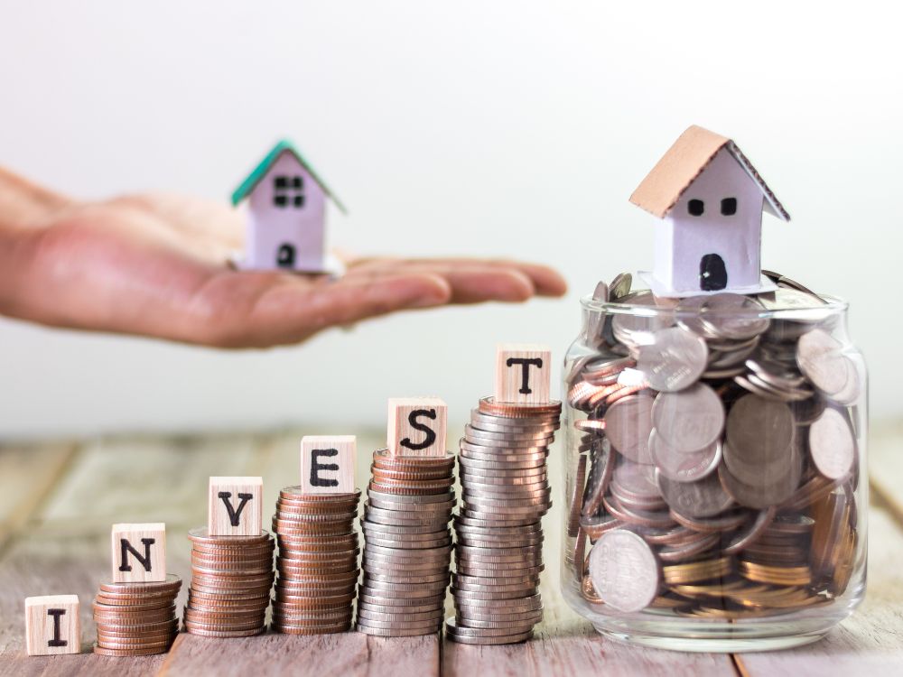 Moedas empilhadas formando a palavra invest ao lado de pote com dinheiro e mini casas, representando investimento imobiliário.
